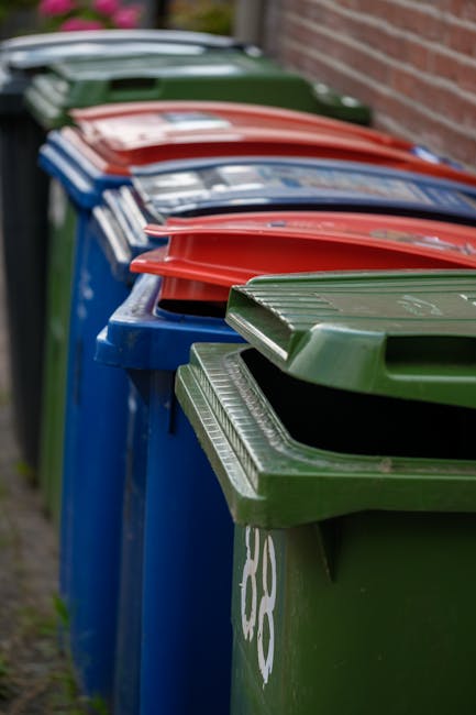 A row of large wheelie bins positioned along a residential sidewalk next to a brick wall, arranged in an alternating pattern of green, blue, red, and dark green. The bins are made of plastic with hinged lids, some of which are open, revealing empty interiors or waste materials. The bins are part of home waste collection services and are typically used for recycling, general waste, and other household rubbish. The image captures the bins standing outdoors on a curbside, with natural daylight illuminating the scene, and the background slightly blurred to focus on the bins. This setup is commonly seen during waste collection or before a move, where clean, organized disposal of household waste is necessary, aligning with professional removals and relocation processes occasionally involved in house removals in West Heath.