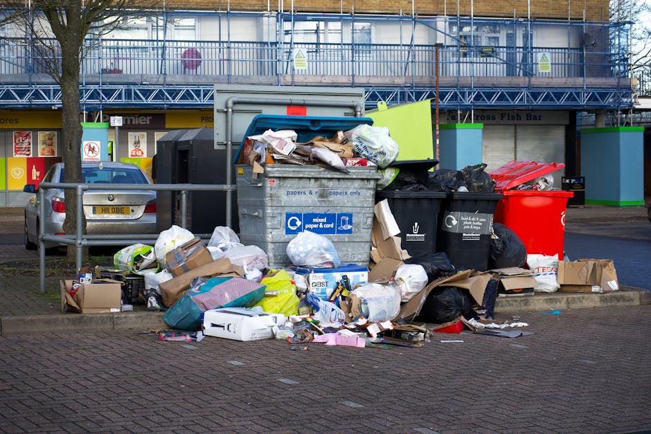 A collection of overflowing domestic waste and recycling bins situated on a paved sidewalk outside residential shops, including a grey mixed waste bin filled with paper, cardboard, and plastic, with its lid open and trash spilling onto the ground. To the left, a black recycling bin is filled with black bags, and a red bin is also visible behind it. Cardboard boxes and packaging materials are scattered around the bins, some flattened or torn, with paper and plastic wrapping among the debris. A grey car is parked close to the waste collection area, partially blocking the view, and a metal railing separates the bins from the pedestrian pathway. In the background, there is a commercial building with shopfronts, advertisements, and a scaffolding-covered upper floor, under an overcast sky. The scene illustrates typical household waste accumulated near a property, relevant to house removals and waste disposal considerations, with natural lighting highlighting the scattered rubbish and urban environment.