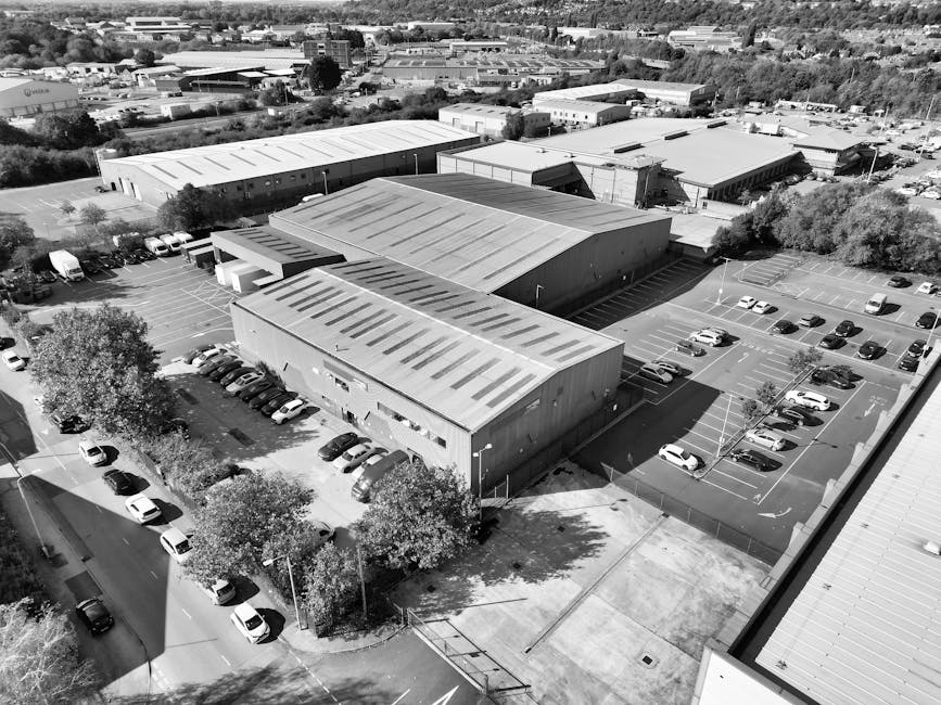 An aerial view of a large industrial estate with multiple metal warehouses featuring flat roofs, some with visible skylights. Surrounding the buildings are arranged parking lots filled with cars, including sedans and vans. There are segregated parking areas; one section is next to a small group of trees and a paved road, while other parking sections are adjacent to the warehouse buildings. In the foreground, a loading area with a few vehicles parked nearby suggests ongoing or recent furniture transport or home relocation activities. The industrial estate is set within a developed commercial and industrial area with additional warehouses and infrastructure visible in the background, under natural daylight conditions that highlight the structures and surroundings. This scene reflects the typical environment where professional removals services, such as those offered by Man with Van West Heath, may operate during large-scale moving or packing and moving efforts.