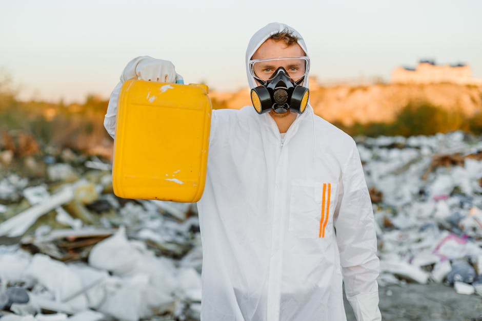 A person wearing a white protective suit, safety goggles, and a black respirator mask is standing outdoors among a large area of scattered waste and debris, including plastic, paper, and discarded materials. The individual is holding a yellow plastic container in their right hand, positioned in front of their body. The background features an open landscape with a distant view of buildings or cliffs under a soft sky. The scene suggests a hazardous waste management or environmental cleanup activity, with the individual prepared for hazardous materials handling. The setting emphasizes the importance of safety gear in waste removal, associated with professional remediation or environmental services, such as those provided by Man with Van West Heath during logistical or home relocation processes involving hazardous waste disposal.