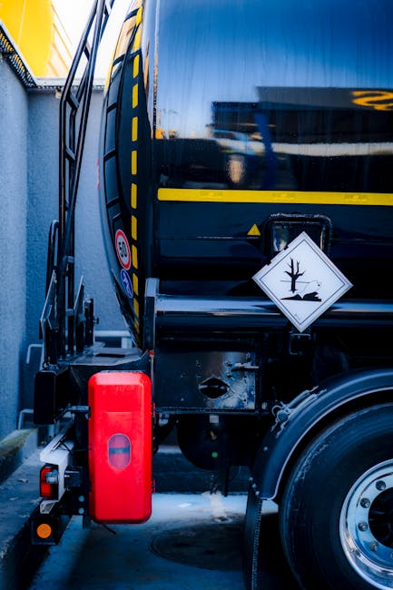 A large black vehicle with yellow hazard markings parked in a narrow outdoor area next to a building, used for home relocation and moving services by Man with Van West Heath. Attached to the vehicle is a red hazardous waste containment box, secured on the side, with a white warning sign displaying a tree with a line through it, indicating hazardous material. The photo captures part of the vehicle's side, including a reflection of the surrounding environment on its surface, and shows the front wheel extending into the frame. The setting appears to be a loading or unloading area, with natural daylight illuminating the scene. This image illustrates the careful handling and transportation of potentially hazardous waste during house removals or relocation processes by professionals experienced in packing and moving tasks. Man with Van West Heath ensures compliance with waste removal regulations for safe and efficient property clearance.