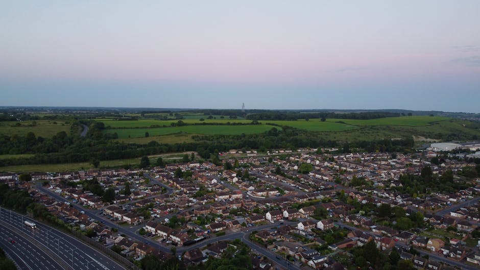 An aerial view of a residential neighbourhood in West Heath during twilight, showing rows of terraced and semi-detached houses with tiled roofs, situated along a network of paved streets lined with parked cars. To the left, a large motorway with multiple lanes and vehicles clearly visible runs parallel to the houses, separated by a buffer zone. Beyond the urban area, expansive green fields, patches of trees, and rolling countryside extend into the distance under a sky transitioning from pale pink to light blue, indicating early evening. This image captures the typical setting for house removals and home relocation activities, with the focus on the residential area adjacent to the transportation infrastructure. The scene highlights a stable environment suitable for furniture transport and packing during moving services, as managed by Man with Van West Heath, which operates in similar logistical contexts associated with narrow-lane charges for West Heath moves as noted on the specified webpage.