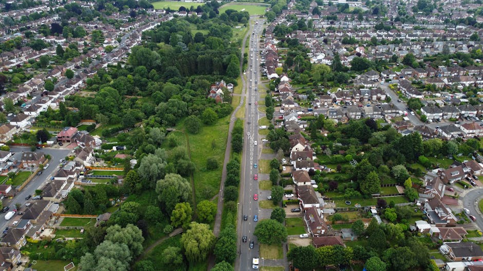 An aerial view of a residential neighbourhood showing a central main road with multiple cars parked along both sides, surrounded by densely packed houses with paved driveways, green lawns, and mature trees. To the left of the road, there is a large green space with a variety of tall trees and open grass areas. On the right, rows of terraced and detached houses with gardens and pathways are visible, interspersed with trees and shrubs. A service vehicle, possibly a van from Man with Van West Heath, is parked on the pavement near the entrance of one property, indicating a home relocation or furniture transport process. The lighting suggests daytime with natural daylight illuminating the scene, which captures the typical environment for local house removals and moving logistics in West Heath.