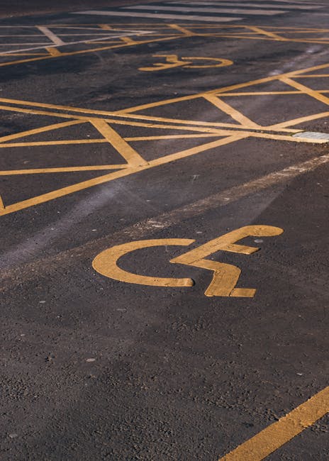 A designated accessible parking space in an outdoor car park, featuring yellow painted lines and a large wheelchair symbol marked on dark asphalt. The space is empty, with no vehicles present, and the painted lines are clearly visible. In the background, part of the adjacent parking spaces with similar yellow markings can be seen. The scene is well-lit, with natural daylight illuminating the area. This parking space is relevant to house removals and home relocation services provided by Man with Van West Heath, highlighting the importance of accessible parking for moving day logistics and safe furniture transport.