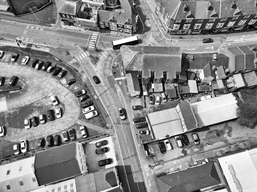 An aerial black-and-white photograph showing a residential area with a street running between buildings and parking areas. In the center, there is a large house with a pitched roof and small garage or storage units attached to its side, surrounded by parked cars on both sides and in nearby parking lots. A white moving truck is parked adjacent to the house, just off the pavement, with several cardboard boxes, wrapped furniture, and packing materials visible nearby, suggesting an active home relocation or furniture transport process. The loading process appears to involve careful placement of items on the driveway, with some boxes and wrapped furniture stacked and being moved to or from the vehicle. Surrounding streets feature additional parked cars and nearby commercial or residential buildings, with clear road markings and traffic signs visible. The overall scene indicates a typical urban move involving organized loading procedures, supported by a professional removal service such as Man with Van West Heath.