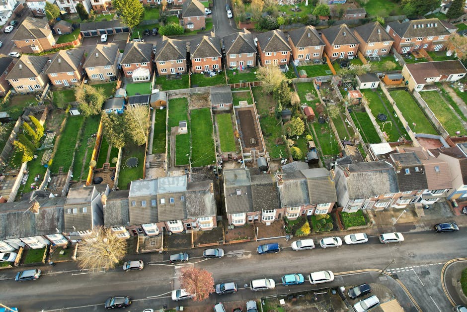 An aerial view of a residential street showing a row of terraced houses with front gardens and pavements. Behind the houses, there are numerous detached and semi-detached houses with fenced back gardens featuring lawns, trees, and garden sheds. Some gardens contain outdoor furniture, children's play equipment, or decorative elements. The roofs of the houses are primarily tiled, with some having solar panels or skylights. Several cars are parked along the street in front of the houses, while others are parked in driveways or within the private gardens. The scene captures a typical suburban neighbourhood, with a focus on layout and property boundaries, emphasizing the context of home relocation and the planning involved with house removals. Occasionally, visible elements such as wheel trolleys, packing materials, or boxes in one or more gardens reflect the process of packing and moving in progress, supporting professional removals by Man with Van West Heath for restricted-access properties and house relocations.