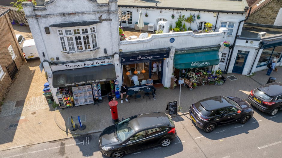 An aerial view of a typical UK high street with three small retail shops positioned side by side. The first shop on the left is a convenience store named Thames Food & Wine, featuring a white facade, a large window display with various snack and beverage items, and a small display stand outside with colorful supermarket shopping baskets. In front of this shop, there is a red pillar post box and two blue bicycle racks. The middle shop, named La Em TRe, has a dark green entrance with a large window, and a sidewalk seating area with three people sitting at a small round table, possibly waiting or taking a break. To the right, there is a flower shop called the Secret Garden, with a green awning, crates and racks displaying an assortment of potted plants, flowers, and garden ornaments. The street in front of the shops has three parked cars, including a black SUV, and a driver is visible walking past or entering one of the shops. The scene indicates a quiet, urban shopping area with pedestrian and vehicle activity, supporting the context of local house removals or moving services conducted nearby by Man with Van West Heath.