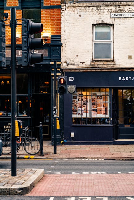 A street scene outside a building on West Heath High Street, showing the exterior of a shop with large glass windows displaying various items, and a blue awning. To the left, a black man with a van is partially visible, wearing casual clothing and working near the vehicle, which is parked on the roadside. Vehicles are stopped at a pedestrian crossing with a red traffic light illuminated; the crosswalk is marked across the road. The surrounding environment includes a brick sidewalk, a bicycle rack, and street signage indicating the location. The scene captures a typical urban area during daylight, with the focus on logistics related to house removals or furniture transport as part of a moving process influenced by the street's commercial activity.