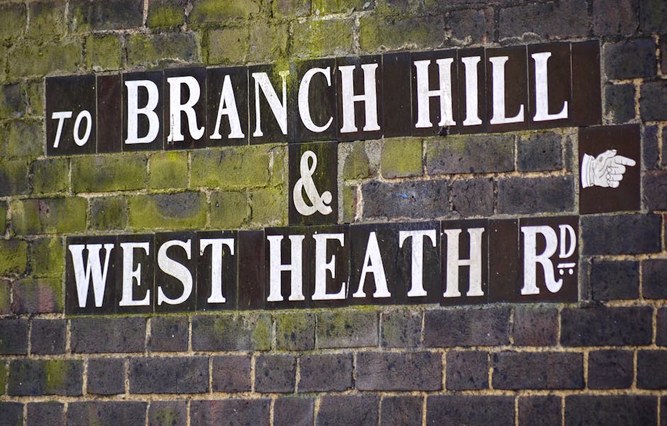 Close-up of a brick wall with moss and weathering, displaying a black and white street name sign reading 'to BRANCH HILL & WEST HEATH R', with a small white pointing hand icon on the right side of the sign, indicating directions. The sign is made of individual rectangular tiles with white lettering on a dark background, affixed to the uneven brick surface. The scene captures the signage typical of residential areas involved in home relocation or moving services, as offered by Man with Van West Heath, situated in an outdoor environment that may be part of a property or street scene related to house removals or furniture transport.