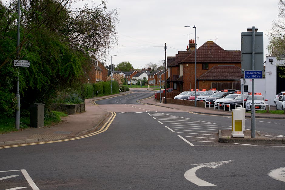 A street scene in West Heath showing a residential area with houses featuring brick facades and pitched roofs. The foreground includes a footpath on the left with a black bin and a lamppost, alongside greenery and trees. There is a parking area on the right, partly sectioned off with white lines, containing multiple parked cars. A sign indicating 'Unsuitable for HGVs' is mounted on a pole near the parking lot. The road has a gentle curve leading away from the foreground, with double yellow lines along the curb. The sky is overcast, and the scene appears to be set during daytime. This setting depicts an environment suitable for home relocation or furniture transport services, with visible parking arrangements, loading zones, and streets suitable for removals companies like Man with Van West Heath.